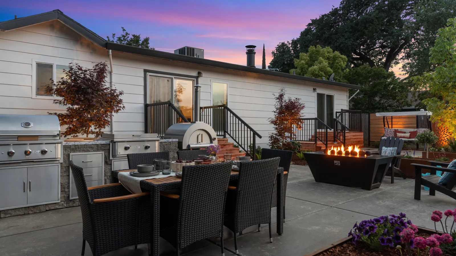 Stainless grill and pizza oven on a stone island beside a gas fire table lounge on a poured patio at dusk, built by Stuteville Landscaping